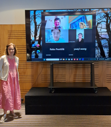 Photo showing woman in pink dress standing next to a screen with online participants
